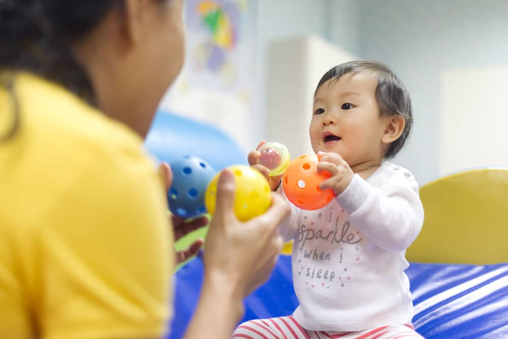 Young little Asian baby holding balls and toys playing with female preschool teacher in classroom